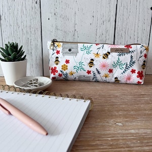 May include: A white pencil case with a zip, decorated with a floral and bee design. The pattern features red, pink, and yellow flowers, green leaves, and cartoon bees. A small brown tag is attached to the side.