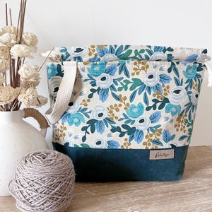May include: A fabric project bag featuring a floral pattern in shades of blue, teal, and mustard yellow on a cream background. The base is a solid dark teal fabric with a small rectangular label. It includes a cream-coloured strap and drawstring.