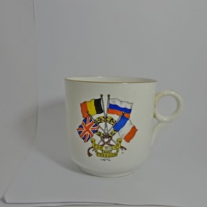 May include: White ceramic mug with a gold rim and a design of four flags crossed together. The flags are the Union Jack, the Belgian flag, the Russian flag, and the French flag. The text "For Right and Freedom" is printed below the flags.