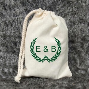 Könnte beinhalten: Eine weiße Baumwolltasche mit Kordelzug, verziert mit einem grünen Lorbeerkranz und den Initialen "E & B" in der Mitte.