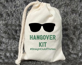 May include: A white drawstring bag with a green and black design. The bag has a pair of sunglasses and the text "Hangover Kit #StraightOutOfThirties".