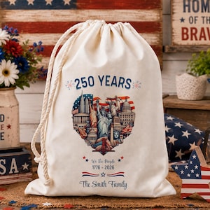 Op de afbeelding: Een crèmekleurige trekkoordtas met de tekst "250 YEARS" en een hartvormige afbeelding met Amerikaanse bezienswaardigheden. De tas bevat ook de tekst "We the People 1776-2026" en "The Smith Family". De achtergrond bevat een Amerikaanse vlag en patriottische decoraties.