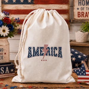 Könnte beinhalten: Ein cremefarbener Kordelzugbeutel mit dem Wort "AMERICA" in blauen, sterngefüllten Buchstaben und einer rot-weiß karierten Blitzgrafik. Die Tasche hat einen Kordelzug aus Seil und steht vor einem patriotischen Hintergrund.