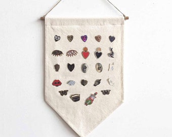Puede incluir: Una pancarta de lona blanca con forma triangular cuelga de una barra de madera. La pancarta está decorada con una variedad de pines coloridos, incluyendo corazones, ojos, calaveras y mariposas.