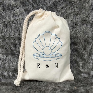 Puede incluir: Una bolsa de algodón blanco con cordón de ajuste, con un contorno azul de una concha y una perla en su interior. La bolsa tiene el texto "R & N" impreso en la parte delantera.