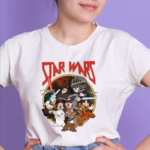May include: White t-shirt featuring a Star Wars graphic with cartoon characters. The design includes the text "STAR WARS" in red, with a circular image of characters like Mickey Mouse, Donald Duck, and Goofy in a space battle scene.