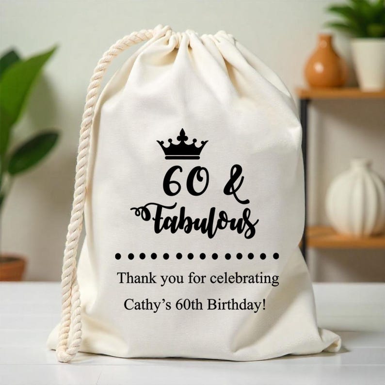 May include: White cotton drawstring bag with black text that reads "60 & Fabulous" with a crown above the text. The bag also has the text "Thank you for celebrating Cathy's 60th Birthday!"