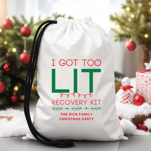 以下が含まれることがあります： 黒い紐が付いた白い巾着袋。赤と緑で「I GOT TOO LIT」と書かれています。その下に「RECOVERY KIT」と「THE NICK FAMILY CHRISTMAS PARTY」が緑色で書かれています。袋はクリスマスツリーの前にあります。