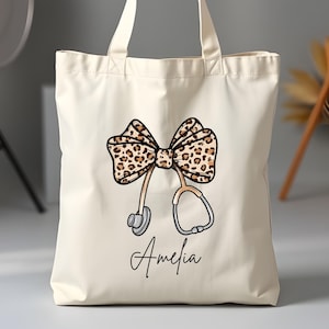 Könnte beinhalten: Eine weiße Canvas-Tragetasche mit einer Schleife im Leopardenmuster und einem Stethoskop. Die Tasche ist mit dem Namen "Amelia" personalisiert.