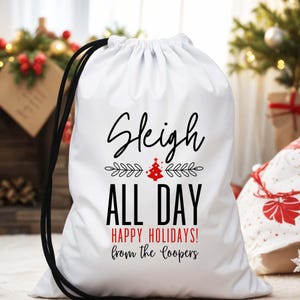 May include: White drawstring gift bag with black drawstring and text that reads "Sleigh ALL DAY HAPPY HOLIDAYS! from the Coopers". The text is in black and red. A red Christmas tree is in the center of the bag.