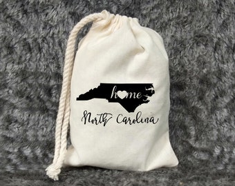 May include: A white drawstring bag with a black outline of the state of North Carolina and the text "home" with a heart in the "o". The text "North Carolina" is written below the state outline.