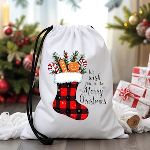 Può includere: Borsa bianca con coulisse con un design a calza natalizia. La calza è a quadri rossi e neri, piena di bastoncini di zucchero, un omino di pan di zenzero e verde. Il testo recita "We wish you a Merry Christmas."