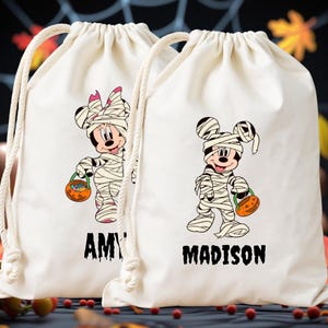 Può includere: Due sacche con coulisse color crema con illustrazioni a tema Halloween. Una sacca presenta Minnie Mouse come mummia, l'altra Topolino. Ogni personaggio tiene in mano un cestino a forma di zucca. I nomi "AMY" e "MADISON" sono stampati sotto.