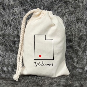 May include: A white drawstring bag with a black outline of the state of Utah and a red heart in the center. The bag has the text "Welcome!" printed on the front.