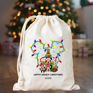 May include: A cream-colored drawstring gift bag featuring a Mickey Mouse head outline made of Christmas lights. Inside the outline are Disney characters, a Christmas tree, and the text "HAPPY DISNEY CHRISTMAS 2025".