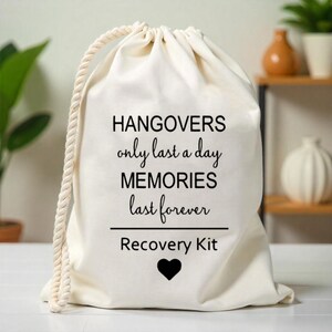 K&ouml;nnte beinhalten: Wei&szlig;e Stofftasche mit Kordelzug und schwarzem Text, der "Hangovers only last a day MEMORIES last forever Recovery Kit" lautet, mit einem schwarzen Herzen unter dem Text.