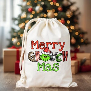 May include: A cream-colored drawstring gift bag with the words "Merry Grinch Mas" in red, green, and black lettering. The Grinch is wearing a Santa hat. The bag is in front of a Christmas tree.