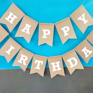 May include: A brown burlap banner with white letters spelling "Happy Birthday". The banner is strung on a blue background.