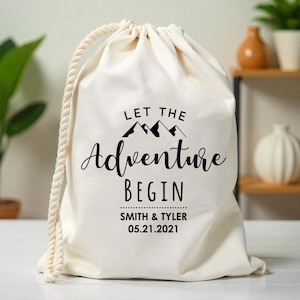 Può includere: Una borsa di cotone bianco con coulisse con il testo "Let the Adventure Begin" e i nomi "Smith & Tyler" e la data "05.21.2021".