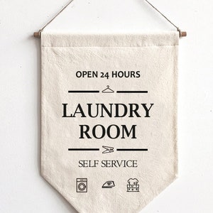 May include: A white canvas banner with black text that reads "Open 24 Hours Laundry Room Self Service". The banner has a hanger at the top and a black and white illustration of a washing machine, iron, and a basket of laundry at the bottom.