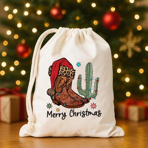 Puede incluir: Una bolsa de regalo con cordón de color crema con un diseño festivo. El diseño incluye botas de vaquero marrones con un gorro de Papá Noel, un cactus con luces navideñas y las palabras "Merry Christmas".