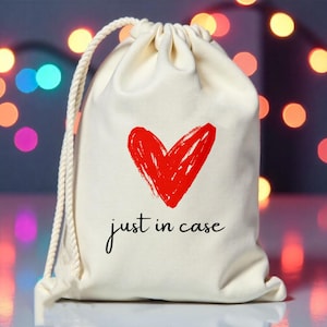 Könnte beinhalten: Eine weiße Kordeltasche mit einem roten Herzen und dem Text "just in case" darauf.
