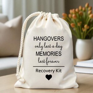 K&ouml;nnte beinhalten: Eine wei&szlig;e Baumwoll-Kordeltasche mit dem Text "Hangovers only last a day MEMORIES last forever Recovery Kit" und einem schwarzen Herzen.