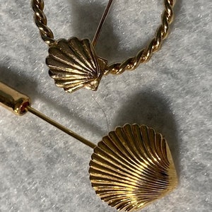 May include: Two gold-toned brooches featuring a seashell design. One brooch is a simple seashell, while the other is a seashell attached to a twisted circle.