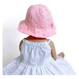 Puede incluir: Un sombrero para el sol de color rosa claro con un dise&ntilde;o floral calado y un peque&ntilde;o lazo. El sombrero lo lleva una persona que viste un vestido blanco. Un accesorio elegante para los d&iacute;as soleados.