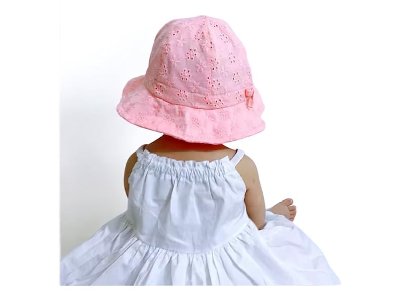 Puede incluir: Un sombrero para el sol de color rosa claro con un dise&ntilde;o floral calado y un peque&ntilde;o lazo. El sombrero lo lleva una persona que viste un vestido blanco. Un accesorio elegante para los d&iacute;as soleados.