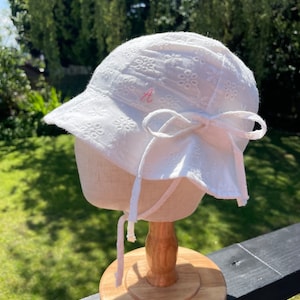 Puede incluir: Sombrero para el sol blanco con un dise&ntilde;o de ojal floral, ala y un lazo grande en la parte trasera. El sombrero tiene una letra "A" bordada en rosa en el lateral y lazos blancos. El sombrero se exhibe en un soporte de madera.