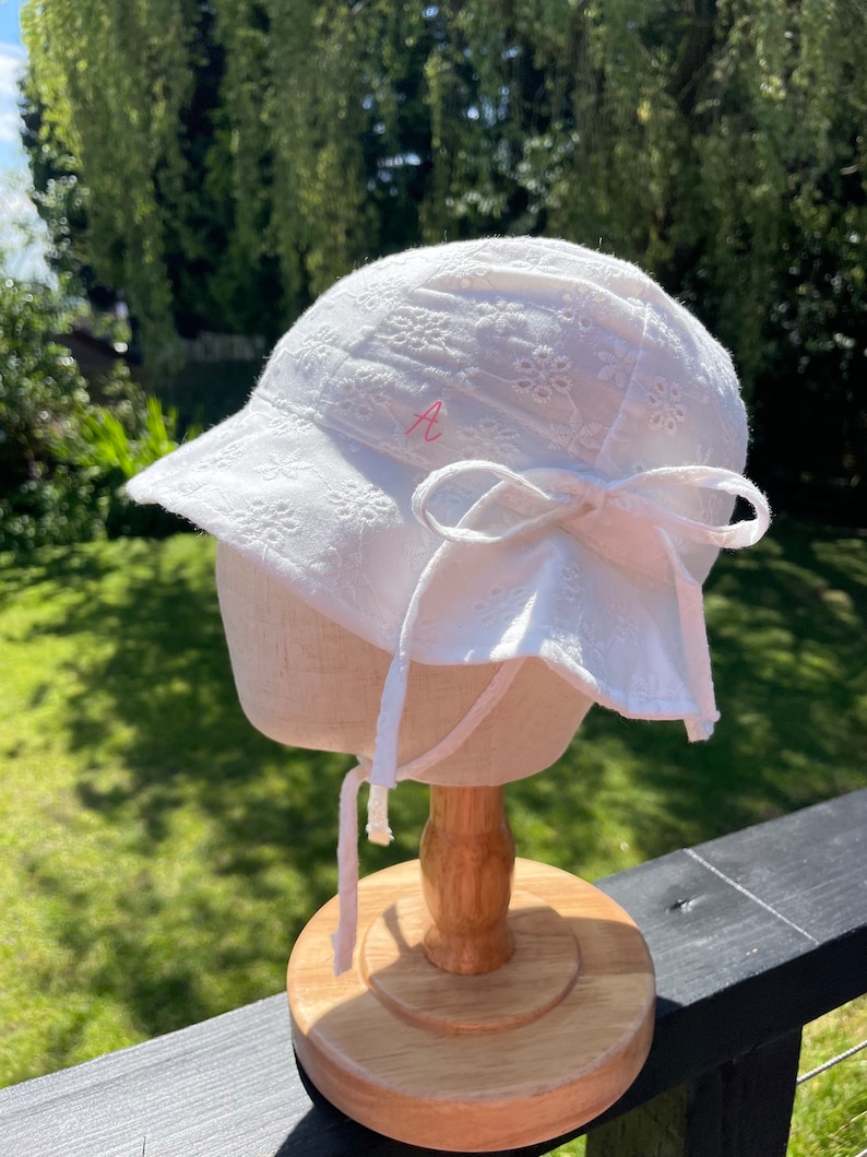 Puede incluir: Sombrero para el sol blanco con un dise&ntilde;o de ojal floral, ala y un lazo grande en la parte trasera. El sombrero tiene una letra "A" bordada en rosa en el lateral y lazos blancos. El sombrero se exhibe en un soporte de madera.