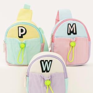 May include: Three colorful sling backpacks in pastel shades of yellow, mint green, pink, and lavender. Each bag features a large letter: "P", "M", and "W". The bags have a front pocket and a neon green zipper pull.