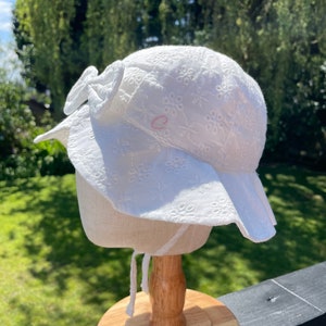 Puede incluir: Sombrero blanco para el sol con ala ancha y lazo. El sombrero presenta bordados florales. Se ve una peque&ntilde;a letra "C" rosa. El sombrero se exhibe en un soporte de madera. Perfecto para los d&iacute;as soleados.