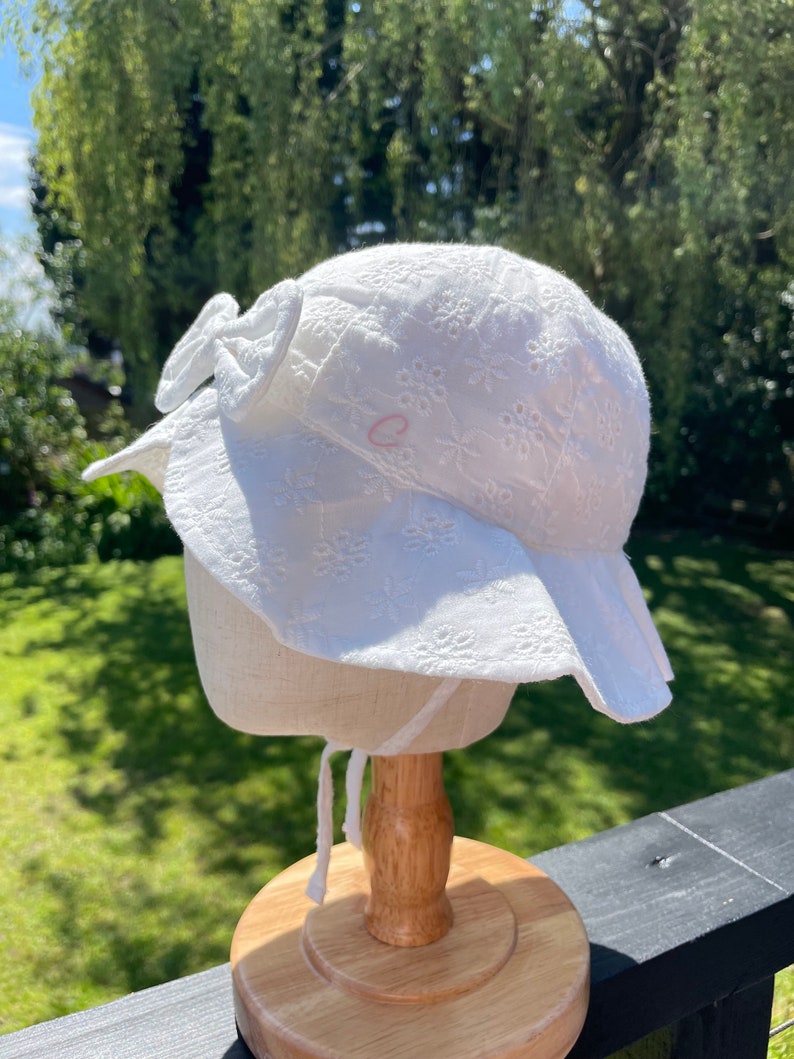 Puede incluir: Sombrero blanco para el sol con ala ancha y lazo. El sombrero presenta bordados florales. Se ve una peque&ntilde;a letra "C" rosa. El sombrero se exhibe en un soporte de madera. Perfecto para los d&iacute;as soleados.