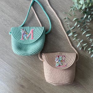 Puede incluir: Dos pequeños bolsos de paja con una correa para el hombro. El bolso de la izquierda es verde con la letra "M" bordada. El bolso de la derecha es beige con la letra "E" bordada.