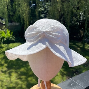 Puede incluir: Un sombrero de sol blanco de encaje ingl&eacute;s con ala ancha y un lazo grande. El sombrero se exhibe en una cabeza de maniqu&iacute;. Presenta un delicado estampado floral y un cord&oacute;n. El fondo es una vista borrosa de follaje verde.