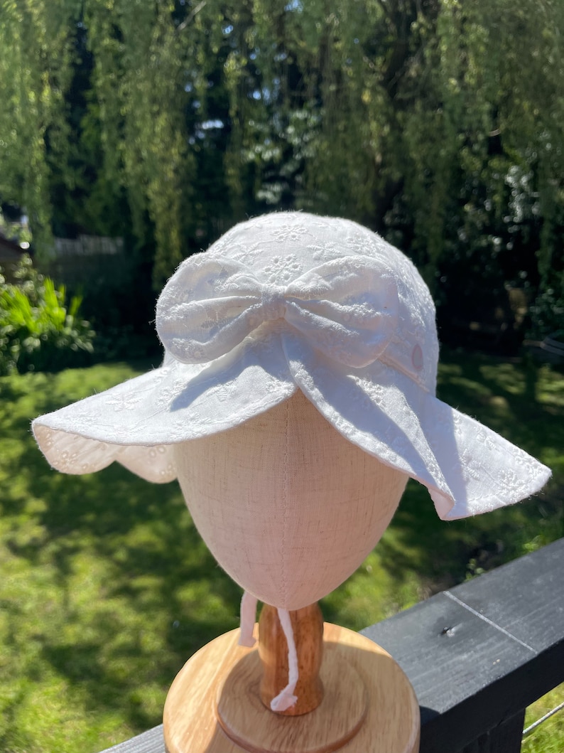 Puede incluir: Un sombrero de sol blanco de encaje ingl&eacute;s con ala ancha y un lazo grande. El sombrero se exhibe en una cabeza de maniqu&iacute;. Presenta un delicado estampado floral y un cord&oacute;n. El fondo es una vista borrosa de follaje verde.