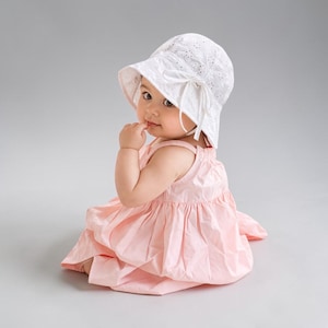 Puede incluir: Un beb&eacute; con un sombrero de sol blanco calado con lazo y un vestido rosa claro. El vestido es sin mangas y tiene una falda fruncida. El beb&eacute; est&aacute; sentado y mirando hacia la c&aacute;mara.