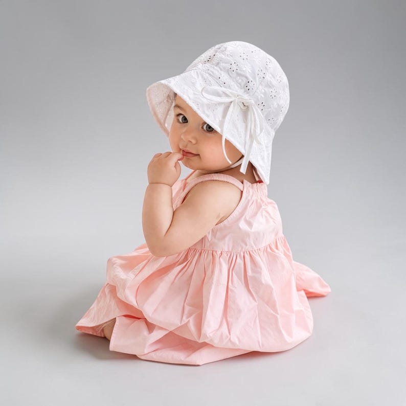 Puede incluir: Un beb&eacute; con un sombrero de sol blanco calado con lazo y un vestido rosa claro. El vestido es sin mangas y tiene una falda fruncida. El beb&eacute; est&aacute; sentado y mirando hacia la c&aacute;mara.