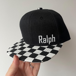 May include: Black baseball cap featuring a chequered peak and the name "Ralph" in white. The cap has a flat peak and a rounded crown. The background is plain white.