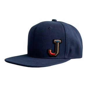 Könnte beinhalten: Marineblaue Baseballkappe mit flachem Schirm. Die Kappe hat einen aufgenähten Aufnäher mit dem Buchstaben "J" in Braun, Rot und Blau. Ein Hut im klassischen Stil, geeignet für Freizeitkleidung.
