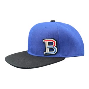Peut inclure: Casquette de baseball bleue avec une visière noire. Le chapeau présente un écusson cousu de la lettre "B" en blanc, rouge et bleu. La casquette a une visière plate et un bouton noir sur le dessus.
