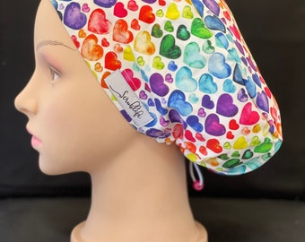 May include: A white scrub hat with a rainbow watercolor heart print. The hat has a drawstring and a label that says "Scrublife".