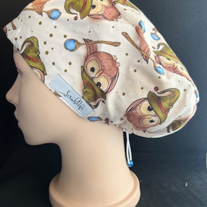 Puede incluir: Un gorro de quirófano blanco con estampado de búhos marrones y verdes. El gorro tiene un cordón de ajuste y una cuenta azul en el extremo del cordón. El gorro tiene la palabra "Scrublife" impresa.