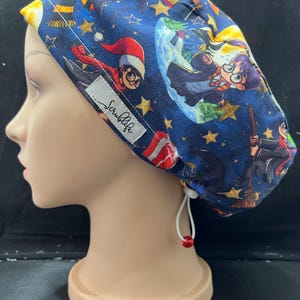 May include: A navy blue scrub cap featuring a repeating pattern of cartoon characters, stars, and other magical elements. The cap includes the text "Scrublife" on a white label. The cap has a white drawstring with a red bead.