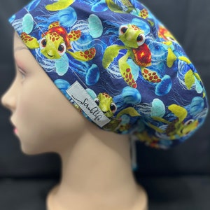 May include: A blue scrub cap featuring a repeating pattern of cartoon sea turtles and jellyfish. The turtles have green and orange shells and are set against a blue background with white wave details. The cap has a white tag that says "Scrublife".