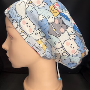 May include: A scrub cap featuring a repeating pattern of cartoon cats and fish in shades of blue, white, and yellow. The fabric cap includes a small grey rectangular tag with the text Scrublife and a white adjustable drawstring with a blue bead.