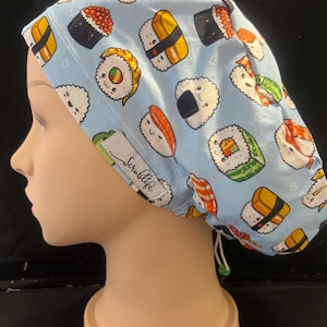 May include: A light blue surgical scrub cap featuring a repeating pattern of various sushi rolls and nigiri. The cap has a drawstring and a small tag that says "Scrublife". The sushi illustrations include salmon, rice balls, and other Japanese food.