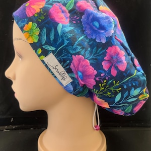 May include: A floral scrub cap with a colourful design of pink, purple, and yellow flowers on a dark blue background. The cap has a white tag that reads "Scrublife".